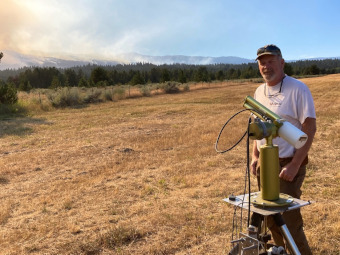 Mr. Mike Arnold with the AERONET sun photometer maintains the Cascade AERONET site.   This photo taken on August 28, 2022 shows smoke from the nearby Four Corners wildfire in the background.   